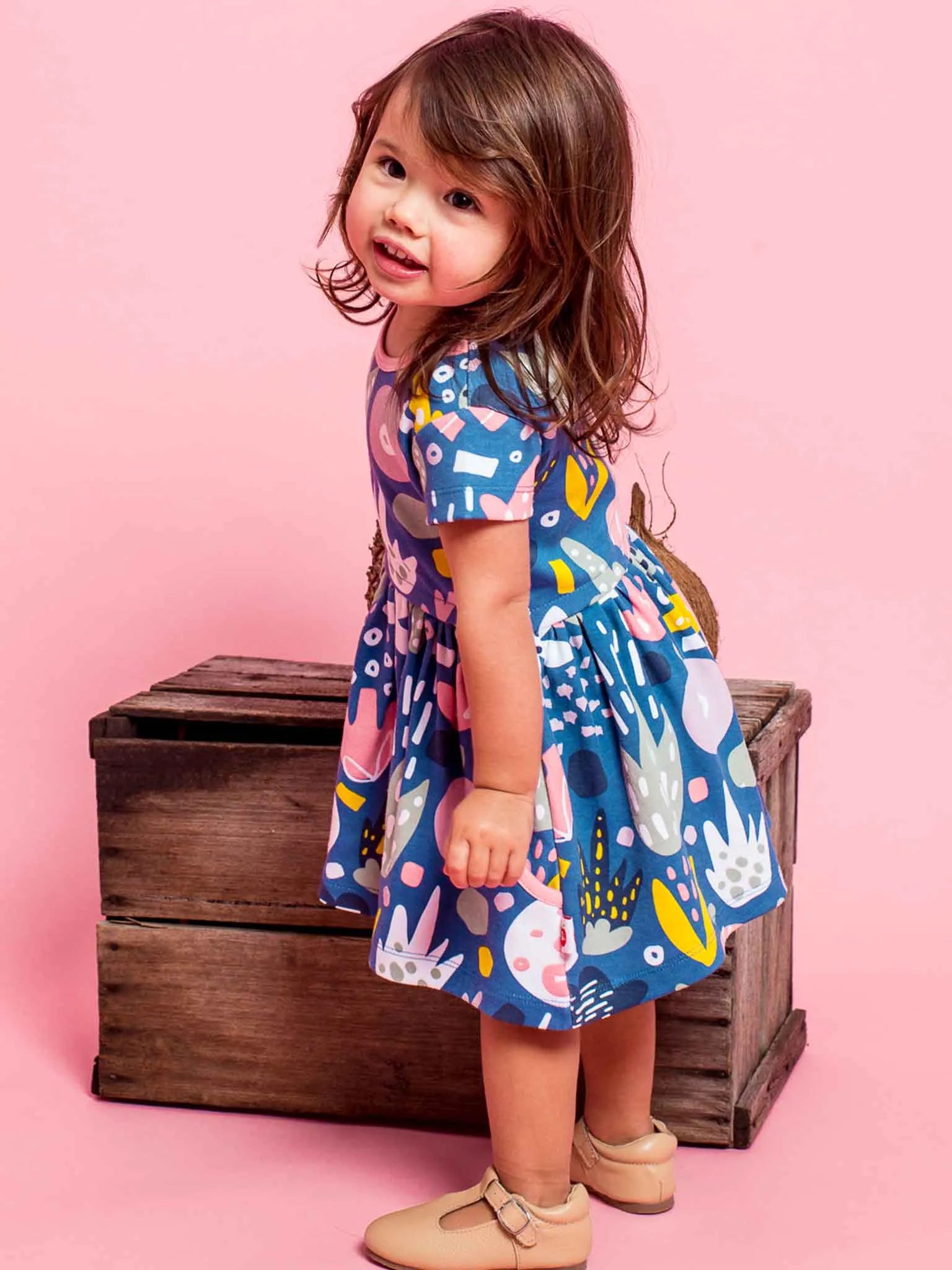 A toddler girl with brown hair stands next to a rustic wooden chest against a solid pink background, wearing a short-sleeve blue dress with a colorful abstract pattern and tan Mary Jane shoes.