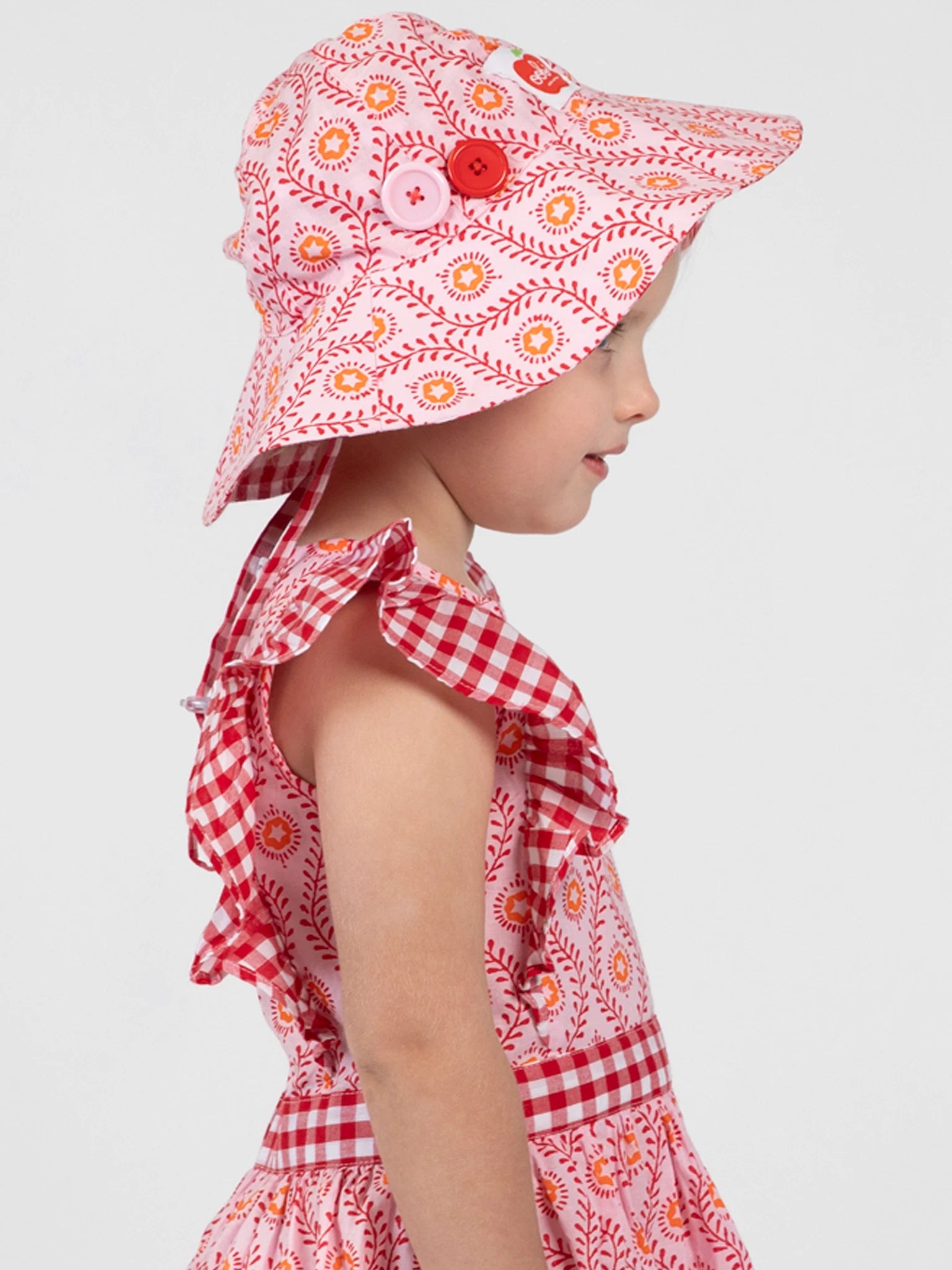 Shown in profile against a plain background, a young child wears the Woodblock Flower Chloe Ponytail Hat by Woodblock Flower with a matching pink and red dress featuring ruffled sleeves.