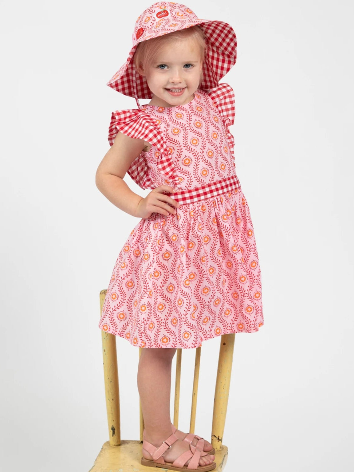 A young girl wears the Woodblock Flower Jayne Dress by Woodblock Flower, featuring a red and white woodblock print, with a matching sunhat and pink sandals, standing on a wooden chair against a plain background.