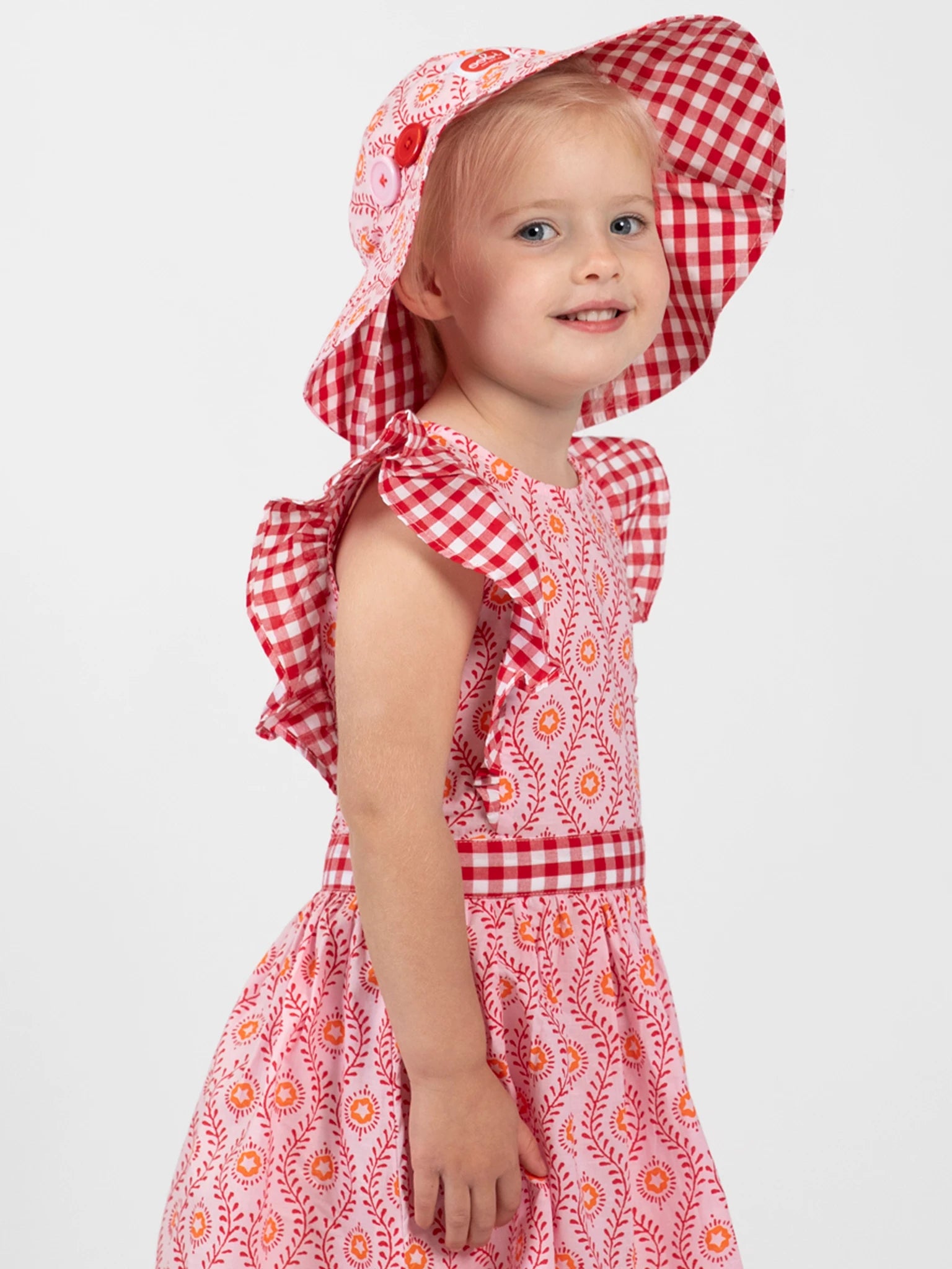 A young child wearing the Woodblock Flower Jayne Dress and a red-and-white checkered sun hat stands smiling against a plain background.
