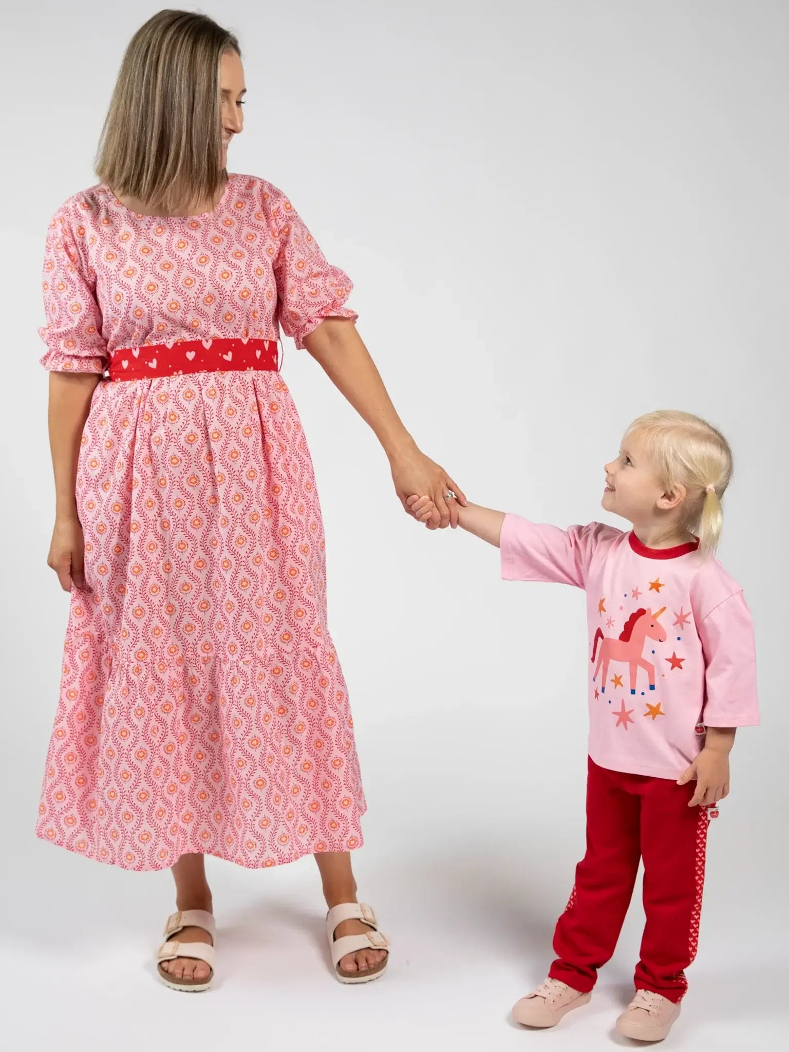 A woman in a Blue Stripe Women's Woodblock Maxi Dress [WH OK] holds hands with a young girl in a pink unicorn shirt and red pants, both standing face-to-face against a plain background.