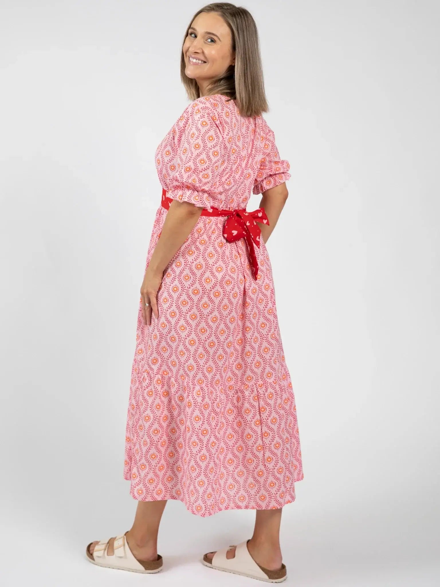A woman stands sideways, smiling, wearing the Blue Stripe Women's Woodblock Maxi Dress [WH OK], featuring a pink pattern, 100% cotton fabric, puff sleeves, red waist tie, and white sandals against a plain white background.
